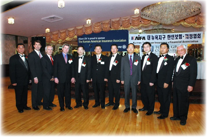A group of men in formal suits and tuxedos standing together at a Korean American Insurance Association event.