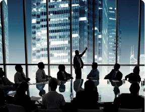 A group of business people sitting in a dark conference room overlooking city skyscrapers.
