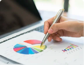 A hand holding a pen pointing at a colorful pie chart on a desk with a laptop.