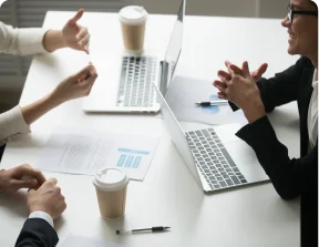 People having a discussion around a white table with laptops and coffee cups.