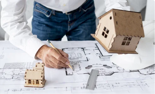 A person drawing on a house blueprint next to miniature wooden house models on a table.
