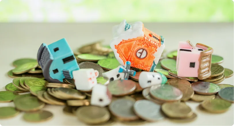 Broken and tipped-over miniature toy houses sitting on top of a large pile of coins, representing property loss or financial risk.