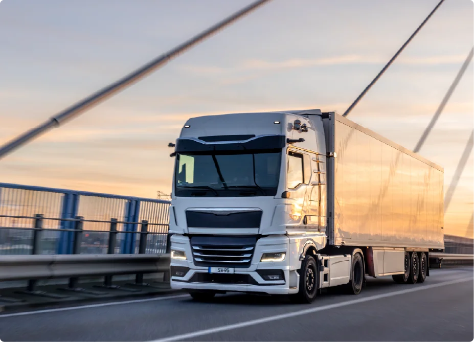 A large white commercial cargo semi-truck driving across a suspension bridge during a bright sunset.