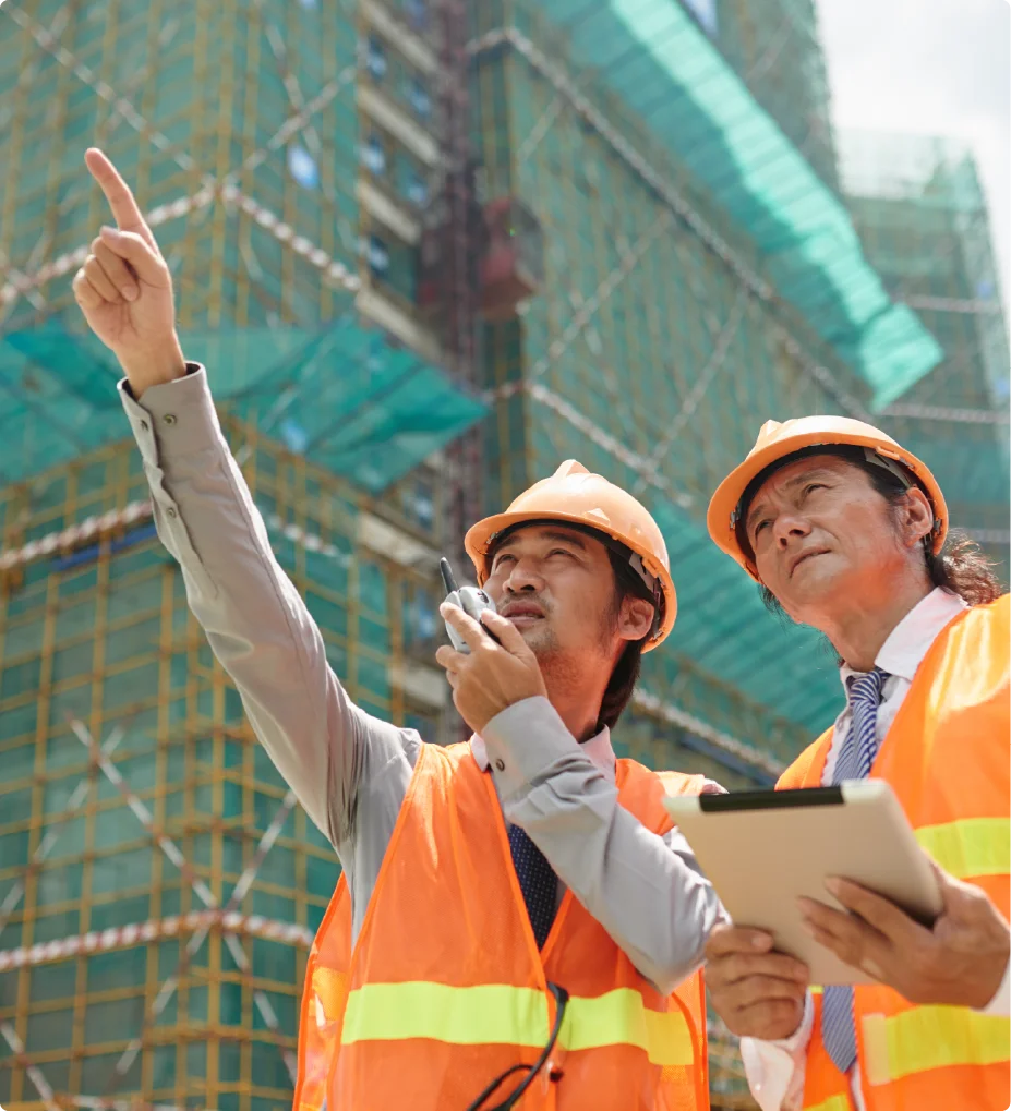 Two construction engineers wearing orange safety vests and hard hats discussing a project at a building site while looking up.