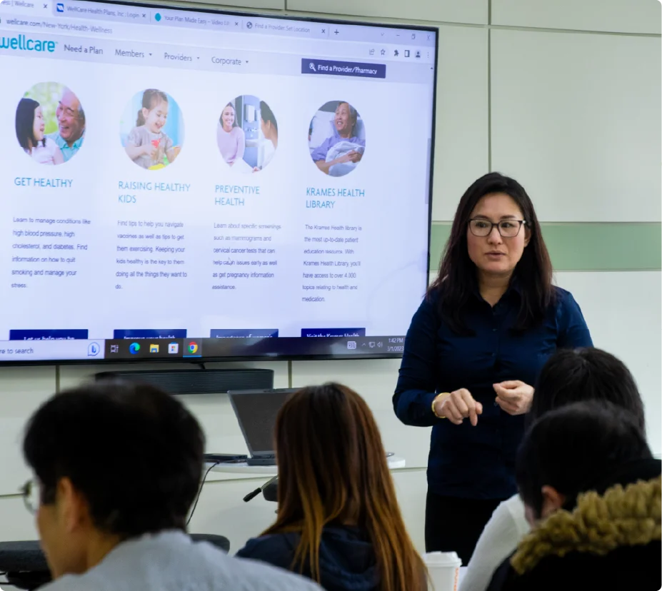 A female professional giving a presentation in front of a large display screen to a group of corporate employees.