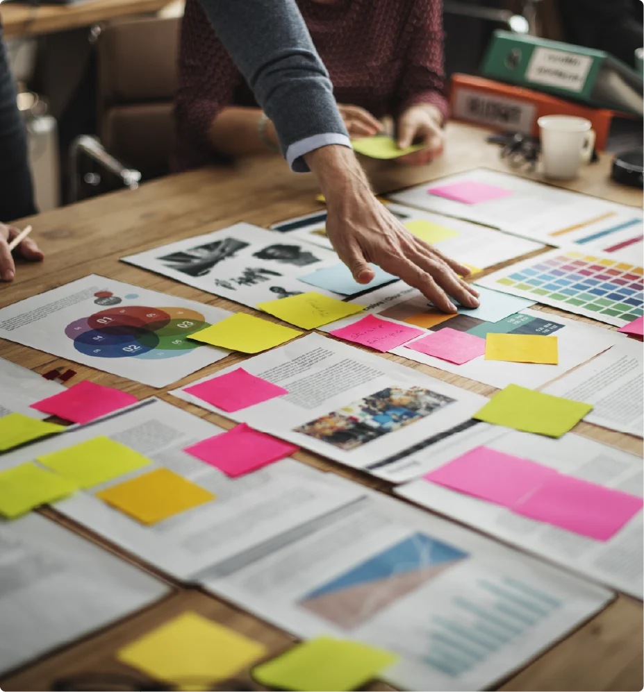 A team collaborating around a wooden desk filled with colorful sticky notes, printed documents, and design mockups.