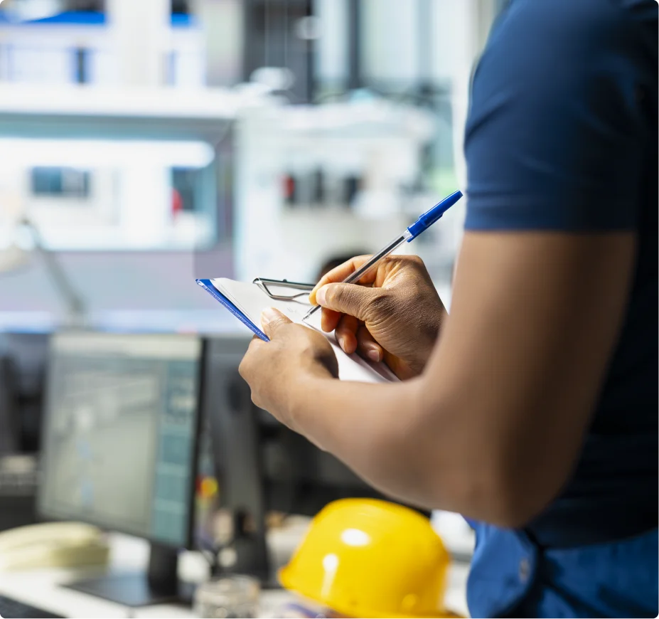 A worker in a blue uniform writing on a clipboard checklist in a factory environment with a yellow hard hat on the table.