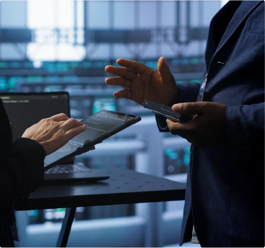 A close-up shot of two IT specialists in a server room, with one person using a tablet and the other holding a hard drive component while discussing data center maintenance.
